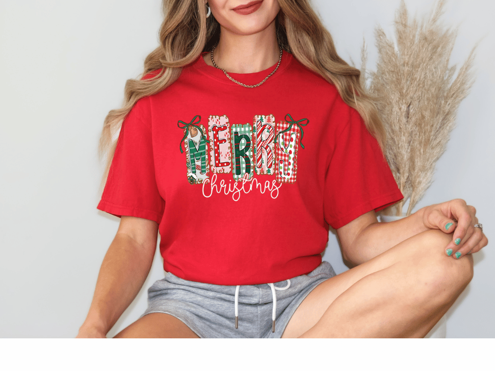 A woman wearing a red t-shirt with the word "MERRY CHRISTMAS" printed on it, sitting with her legs crossed and smiling.