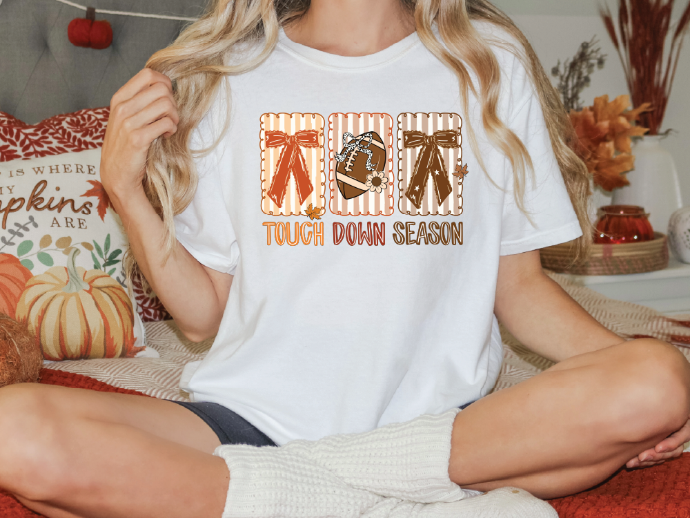 A woman wearing a white t-shirt with a graphic design of a football and the text "TOUGH DOWN SEASON" is sitting on a bed with autumn-themed pillows.