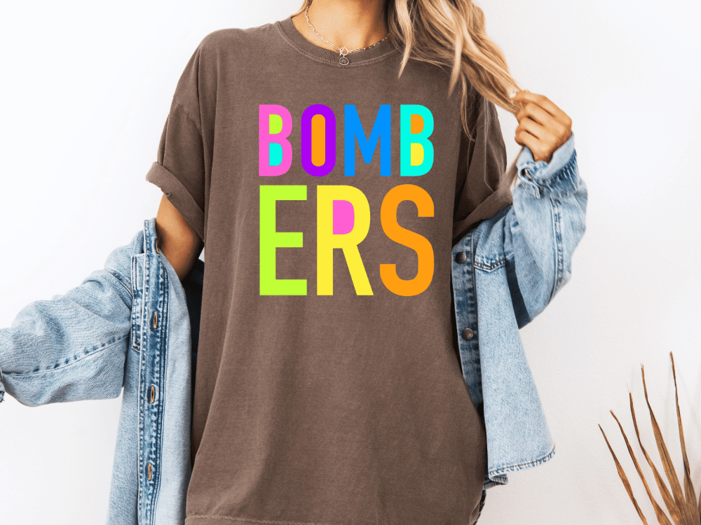 A person wearing a brown t-shirt with the text "BOMBERS" in colorful letters, along with a denim jacket, stands against a white background.