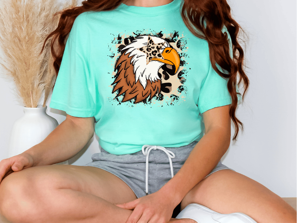 A woman wearing a turquoise t-shirt with a graphic of a leopard print eagle on it, sitting on the floor.