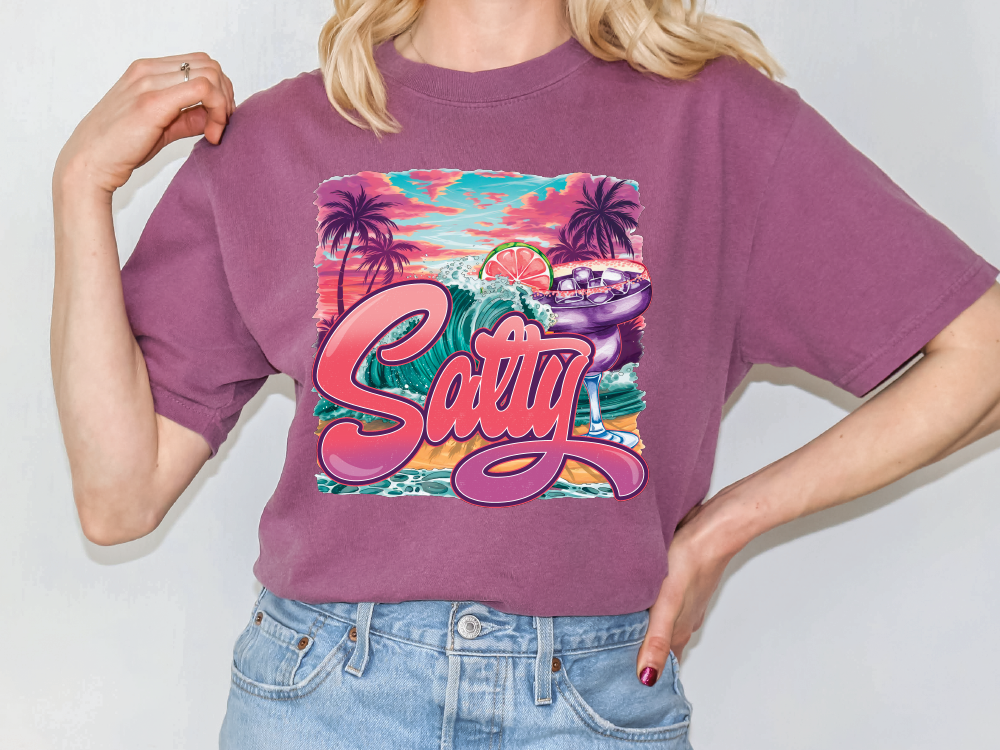 A person wearing a purple t-shirt with a graphic design featuring a beach scene, palm trees, and a surfboard.