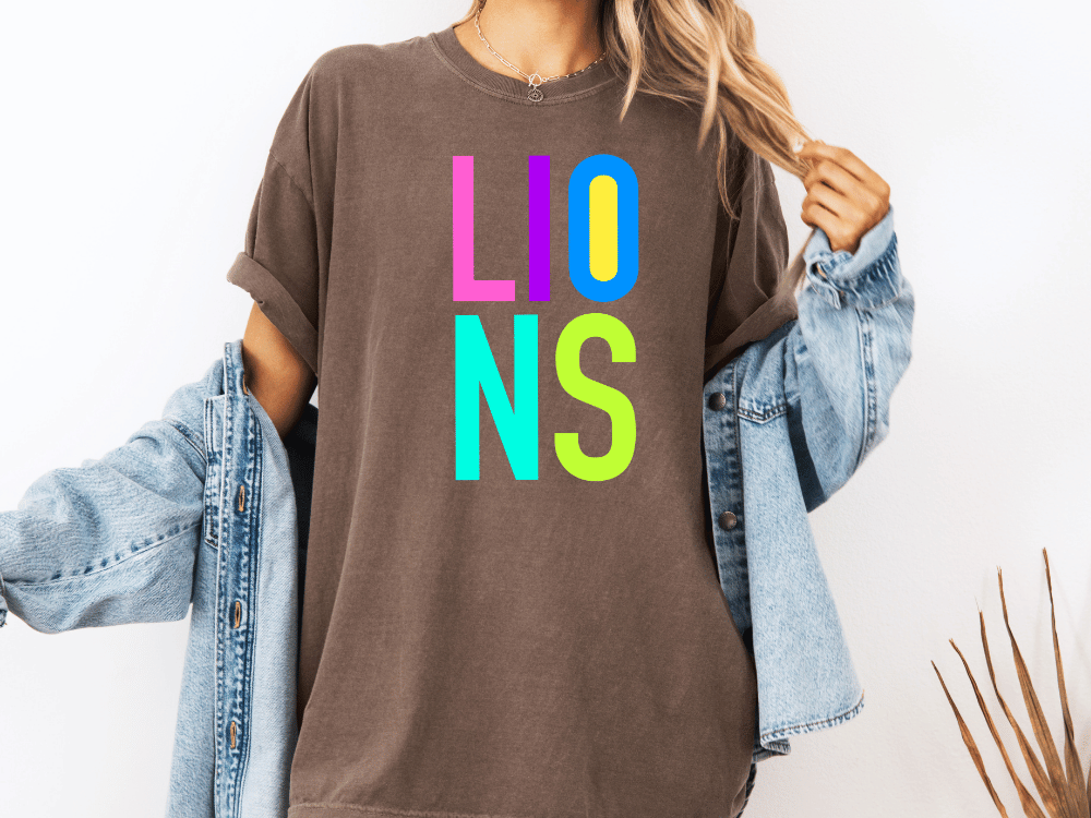 A person wearing a brown t-shirt with the word "LIONS" in colorful letters, along with a denim jacket, stands against a white background.
