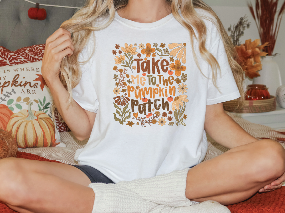 A woman wearing a white t-shirt with a pumpkin patch design and the text "Take me to the pumpkin patch" is sitting on a bed with a pillow featuring a pumpkin and fall-themed design.