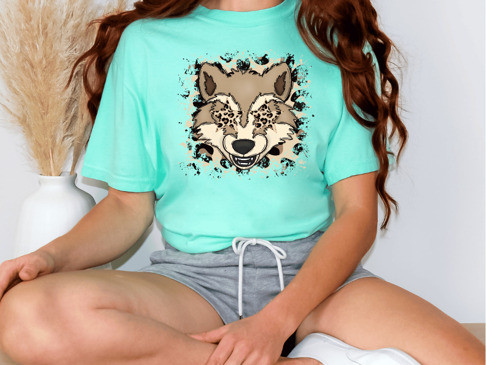A person wearing a turquoise t-shirt with a graphic of a wolf's face and a white skirt is sitting cross-legged on the floor.