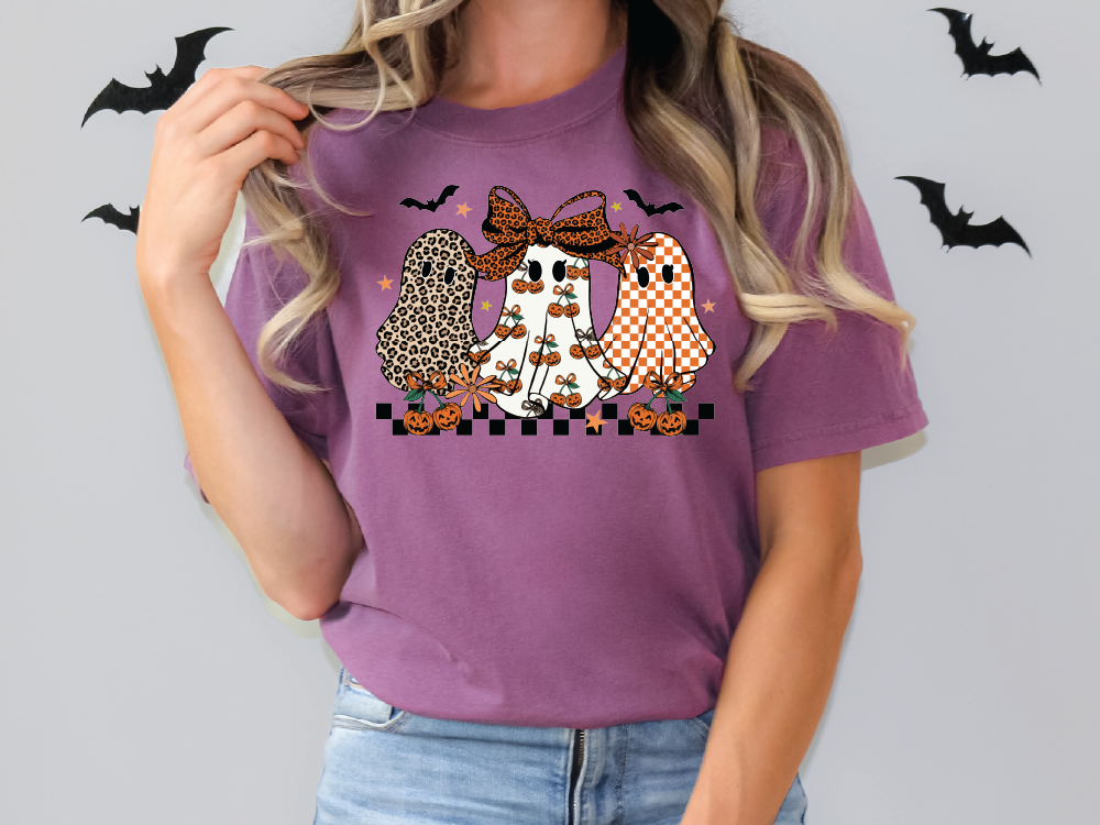 A woman wearing a purple t-shirt with a graphic design of three ghostly figures, including a leopard-print ghost, a checkered ghost, and a pumpkin-wearing ghost.
