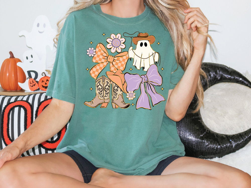 A woman wearing a green t-shirt with a graphic of a ghost, bow, and boots, sitting on a couch with Halloween decorations in the background.