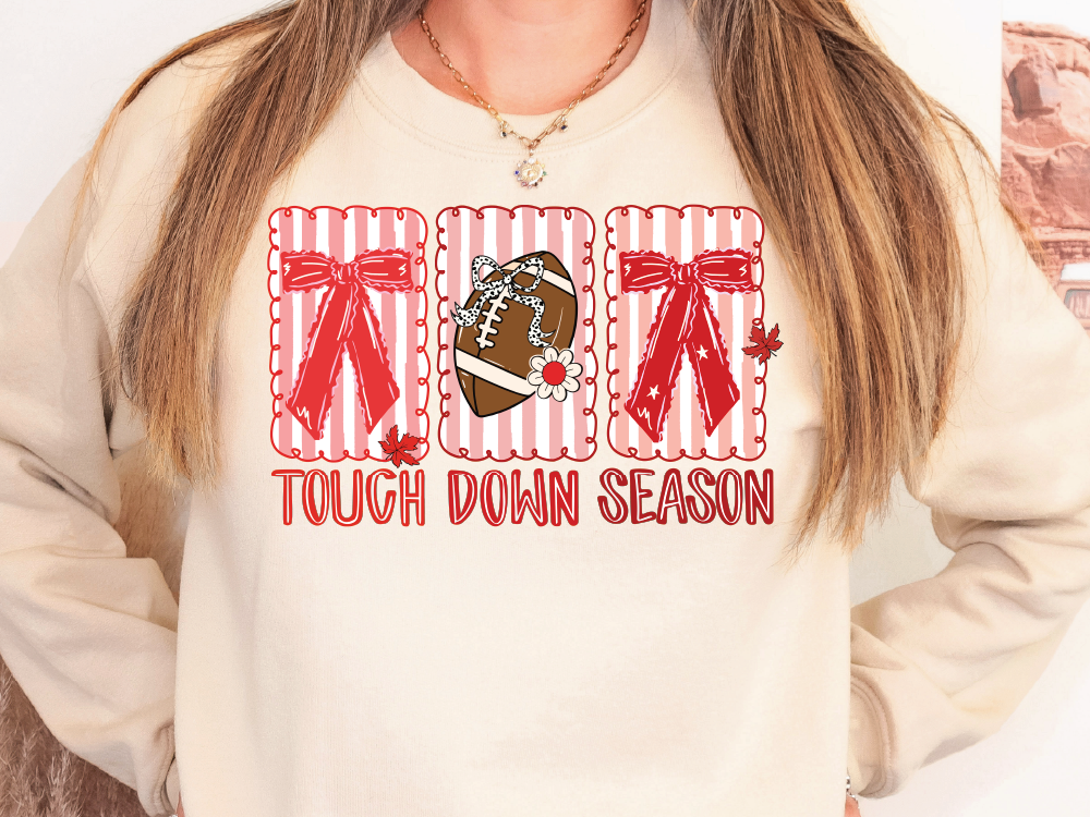 A woman wearing a white sweatshirt with a graphic design featuring a football, red bows, and the text "TOUGH DOWN SEASON".