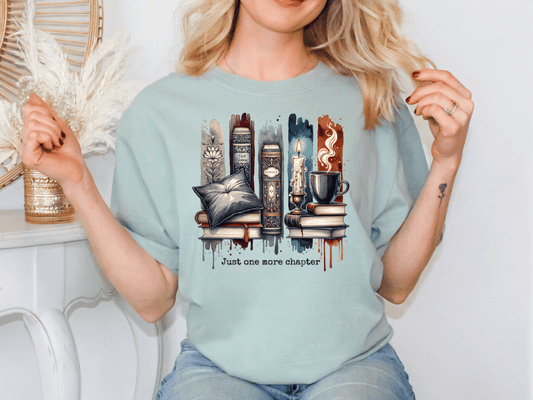 A woman wearing a light blue t-shirt with a graphic design of stacked books and various objects, including a candle, mug, and vase, along with the text "Just one more chapter".