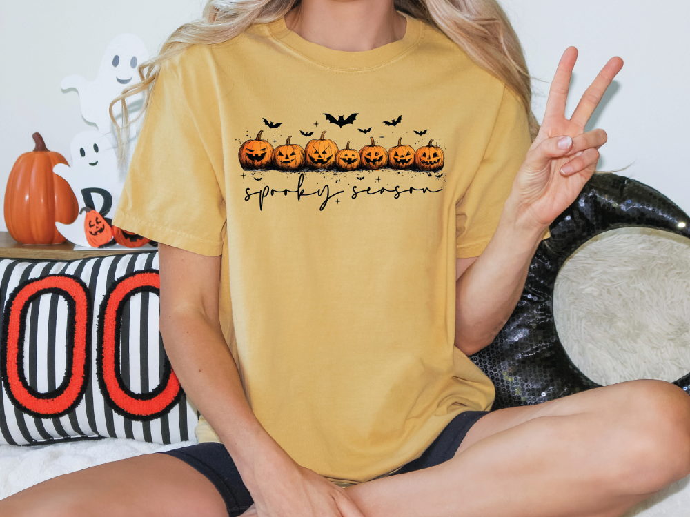 A person is sitting on the floor with a yellow t-shirt featuring a Halloween-themed design, including jack-o-lanterns and bats.