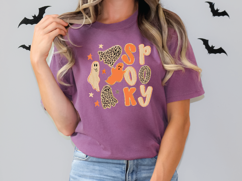 A woman wearing a purple t-shirt with a Halloween-themed design, including a ghost, a leopard print, and the word "Spooky" written on it.