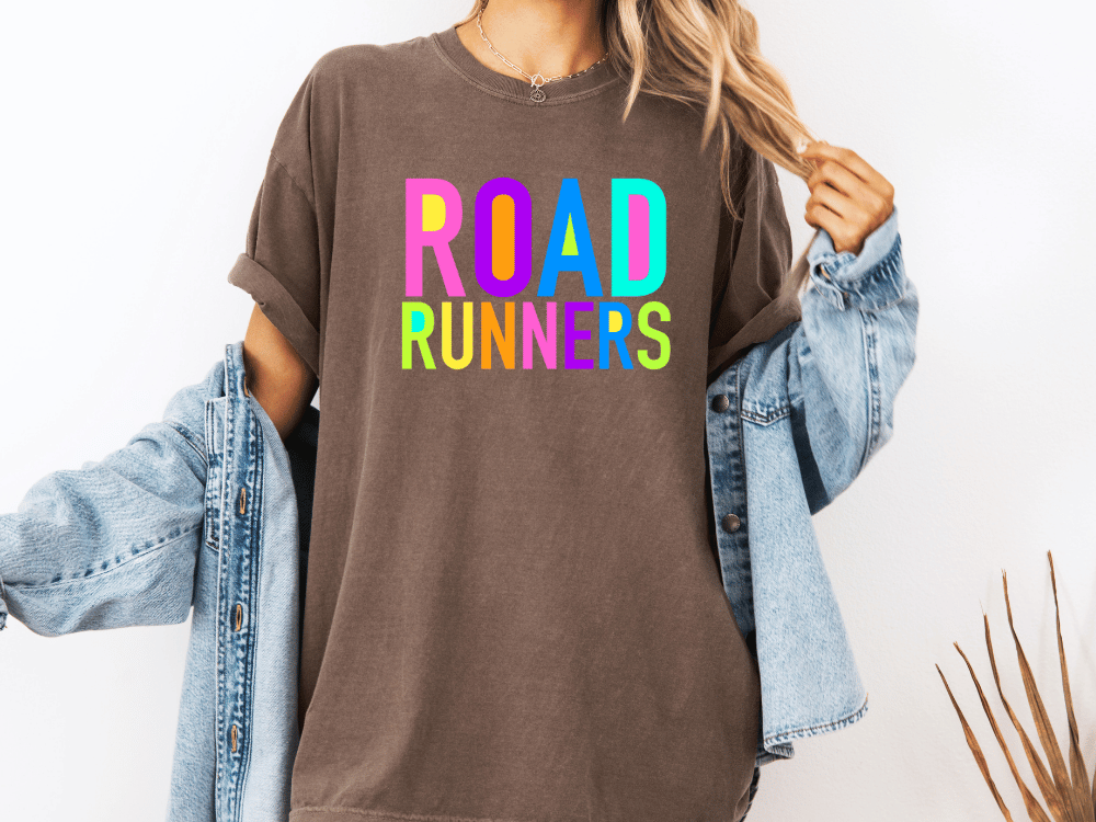 A person wearing a brown t-shirt with the text "ROAD RUNNERS" in colorful letters, along with a denim jacket, stands against a white background.