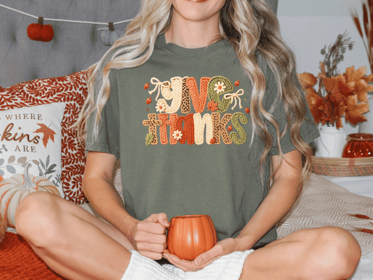 A woman sitting on a bed with a pumpkin-themed pillow and a mug in her hands, wearing a green t-shirt with a Thanksgiving-themed design.