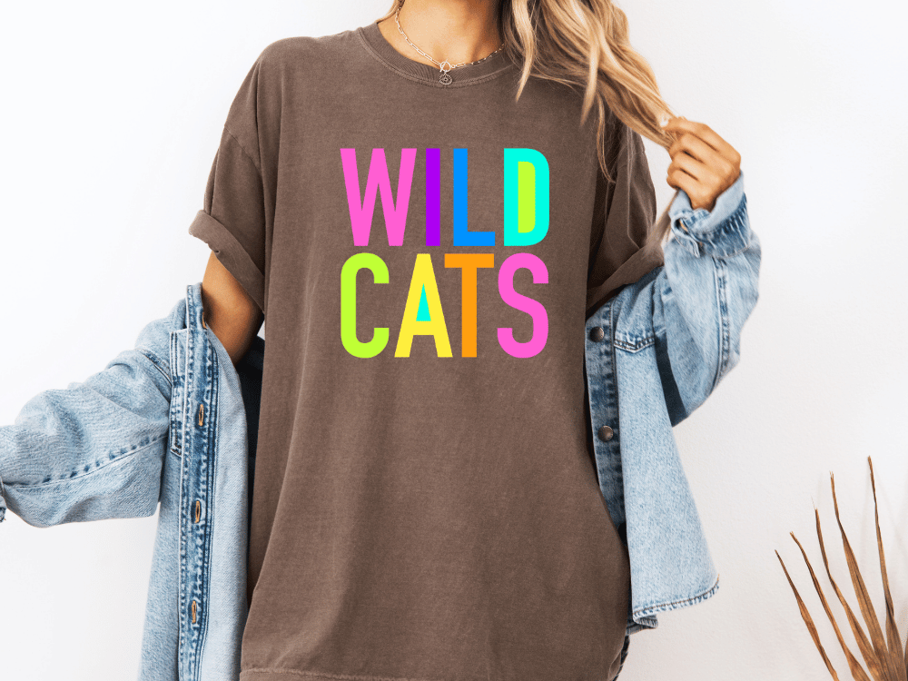 A person wearing a brown t-shirt with the text "WILD CATS" in colorful letters, along with a denim jacket, stands against a white background.