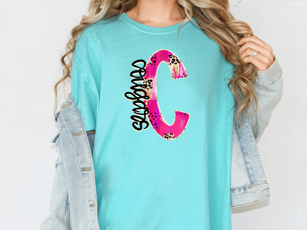 A woman wearing a turquoise t-shirt with a large pink and black letter 'C' printed on it, along with a denim jacket.