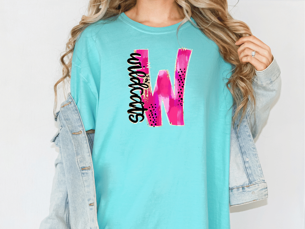 A woman wearing a turquoise t-shirt with a pink and black graphic design on it, and a denim jacket.