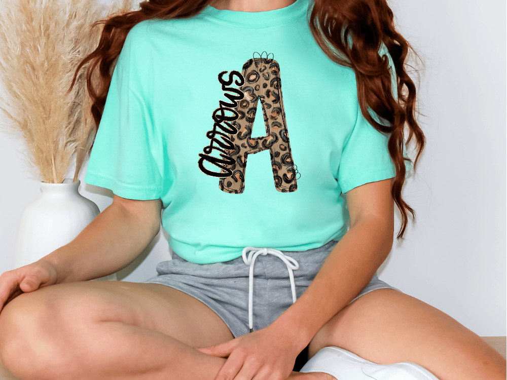 A person wearing a turquoise t-shirt with a leopard print design and gray shorts is sitting on the floor.