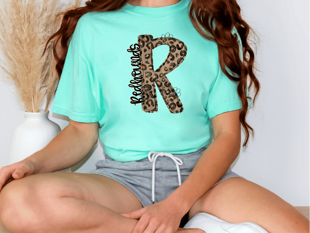 A person wearing a turquoise t-shirt with a leopard print design and gray shorts is sitting on the floor.