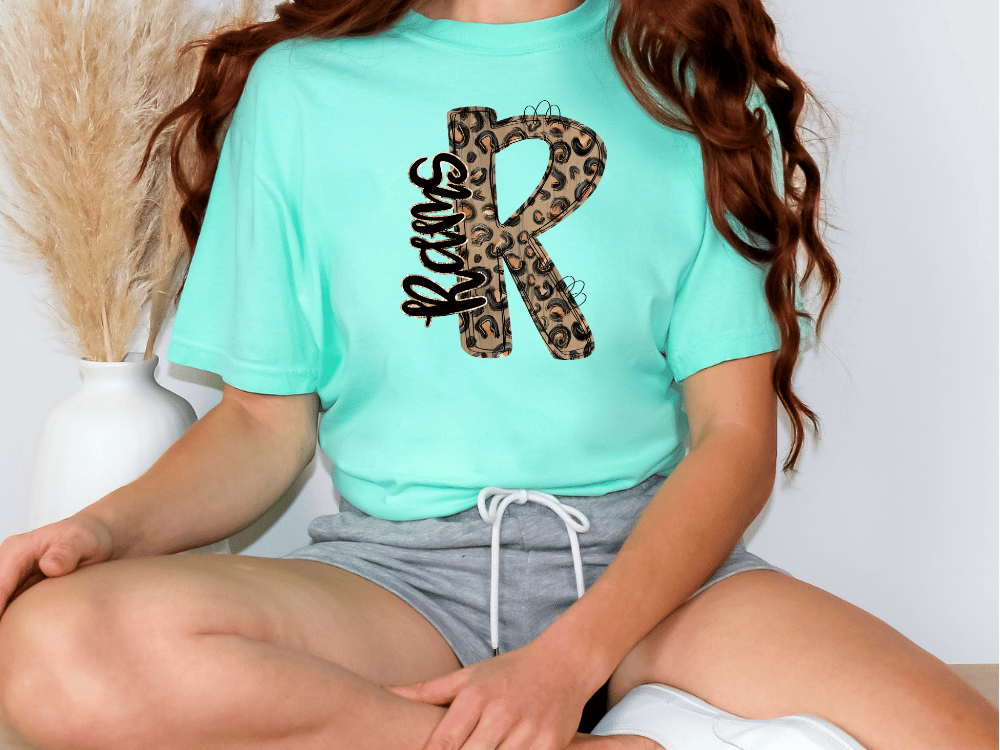 A person wearing a mint green t-shirt with a leopard print design and gray shorts is sitting on the floor.