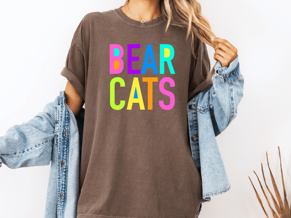 A person wearing a brown t-shirt with the text "BEAR CATS" in colorful letters, along with a denim jacket, stands against a white background.