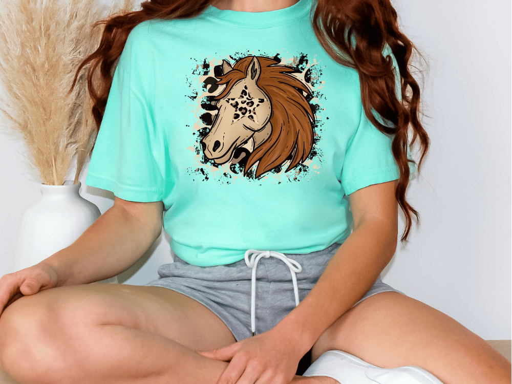 A woman wearing a turquoise t-shirt with a graphic of a horse's head and a patterned design, sitting cross-legged on the floor.