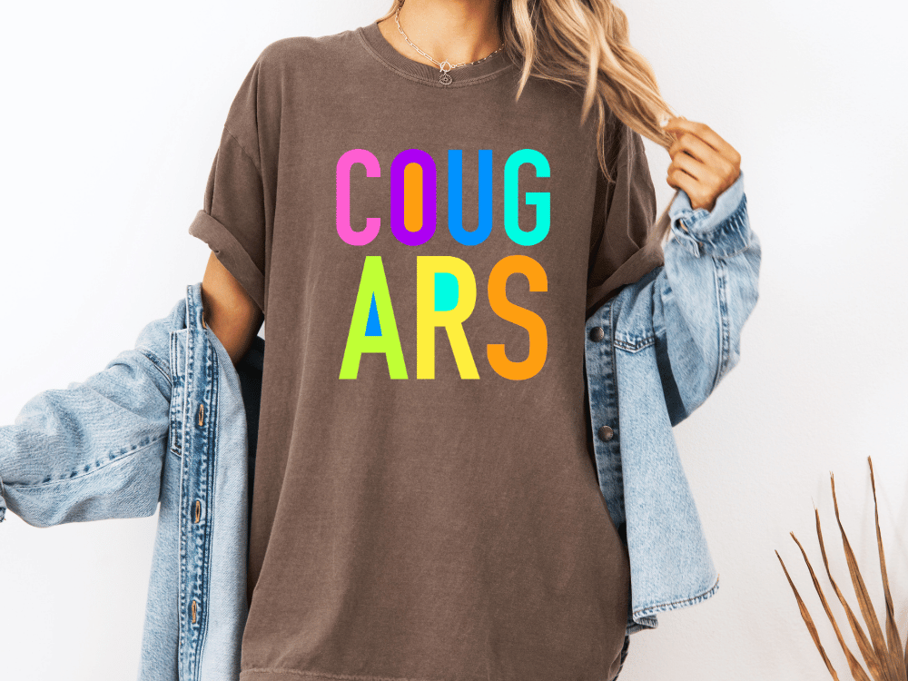 A person wearing a brown t-shirt with the text "COUGARS" in colorful letters, paired with a denim jacket.