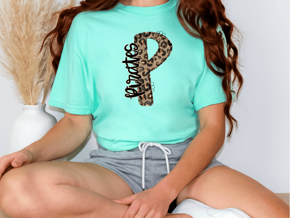 A person wearing a turquoise t-shirt with a leopard print design and gray shorts is sitting on the floor.