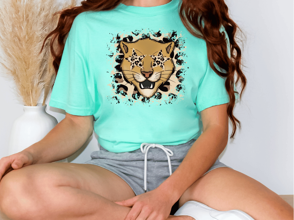 A person wearing a turquoise t-shirt with a leopard print design and gray shorts is sitting cross-legged on the floor.