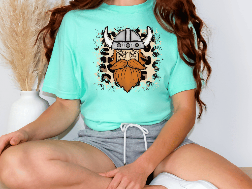 A person wearing a turquoise t-shirt with a graphic of a Viking helmet and beard, sitting cross-legged on the floor.