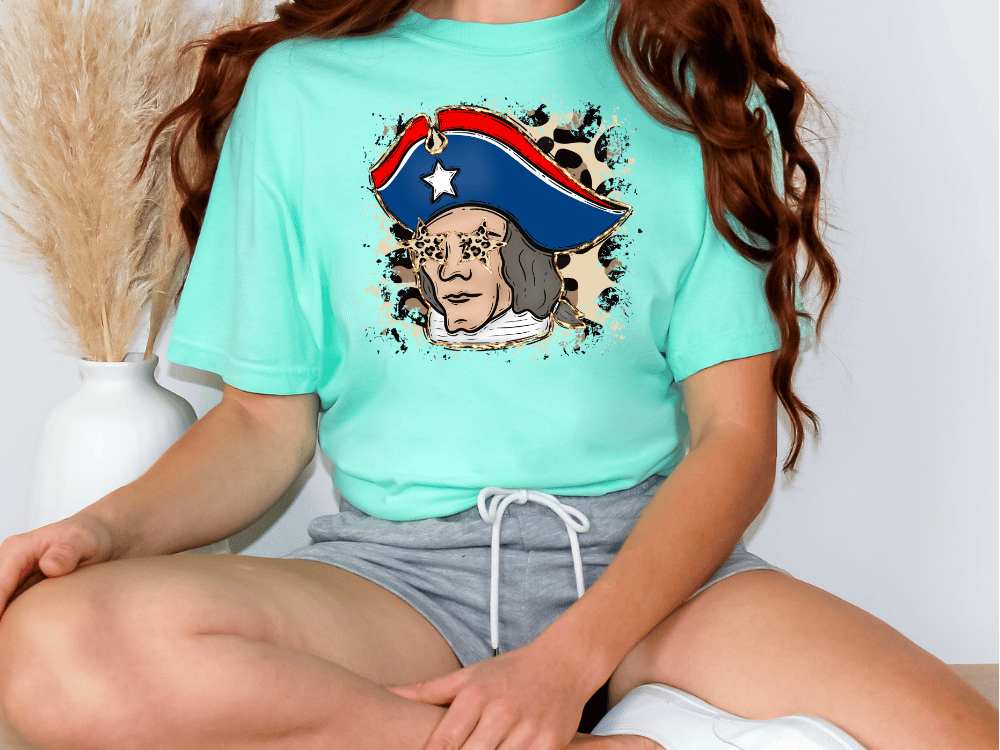 A woman wearing a turquoise t-shirt with a graphic of a man wearing a pirate hat, sitting on the floor.