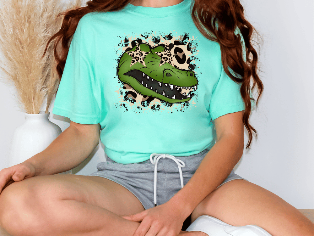 A person wearing a turquoise t-shirt with a graphic of a green alligator and leopard print, sitting cross-legged on the floor.
