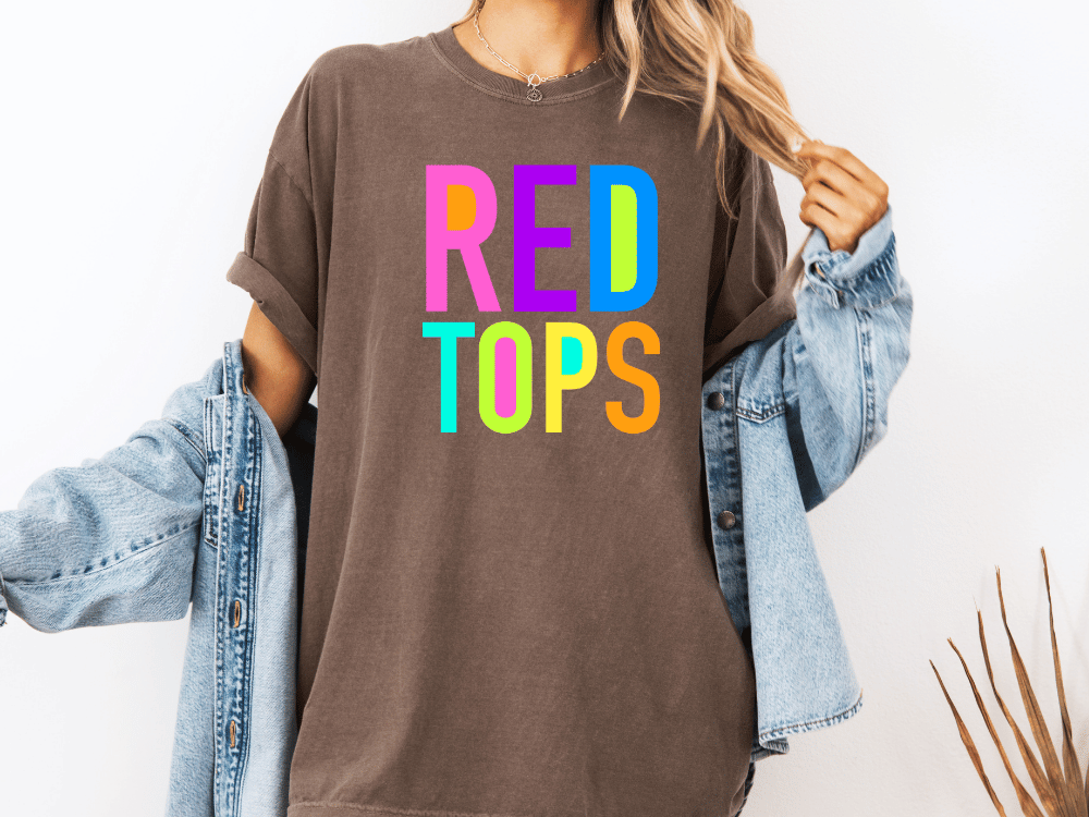 A person wearing a brown t-shirt with the text "RED TOPS" in colorful letters, along with a denim jacket, stands against a white background.