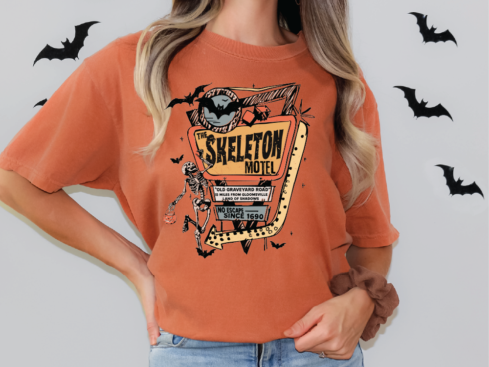 A woman wearing an orange t-shirt with a graphic design of a skeleton motel and bats.