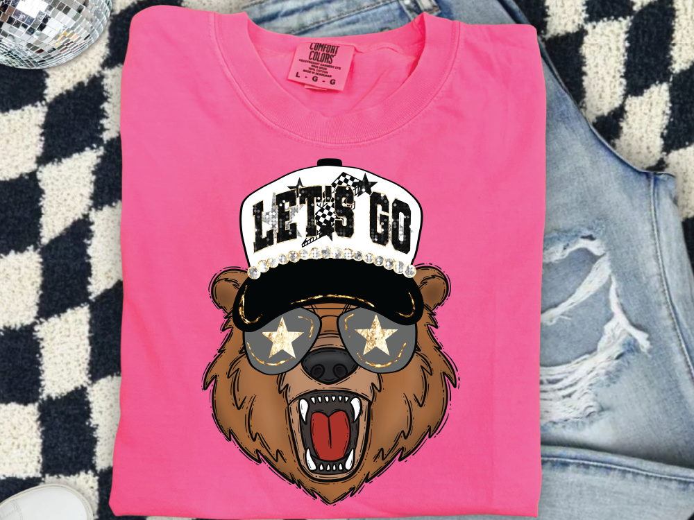 A pink t-shirt with a cartoon bear wearing sunglasses and a hat with a star design, along with the text "LET'S GO" printed on it.