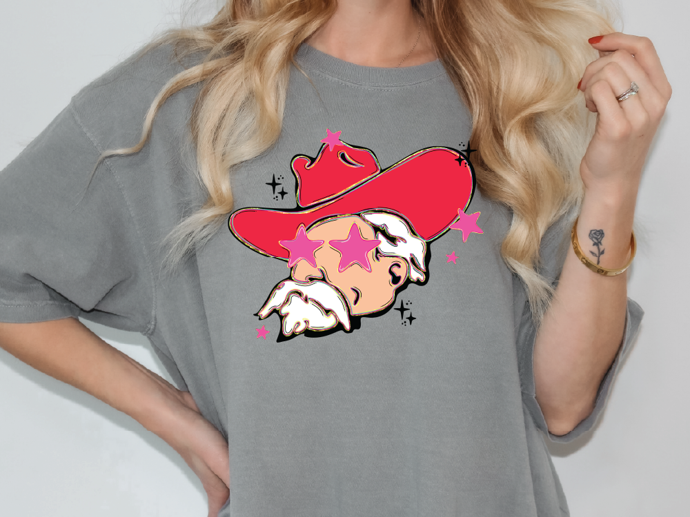 A woman wearing a gray t-shirt with a graphic design of a man wearing a red hat and sunglasses, surrounded by pink stars.