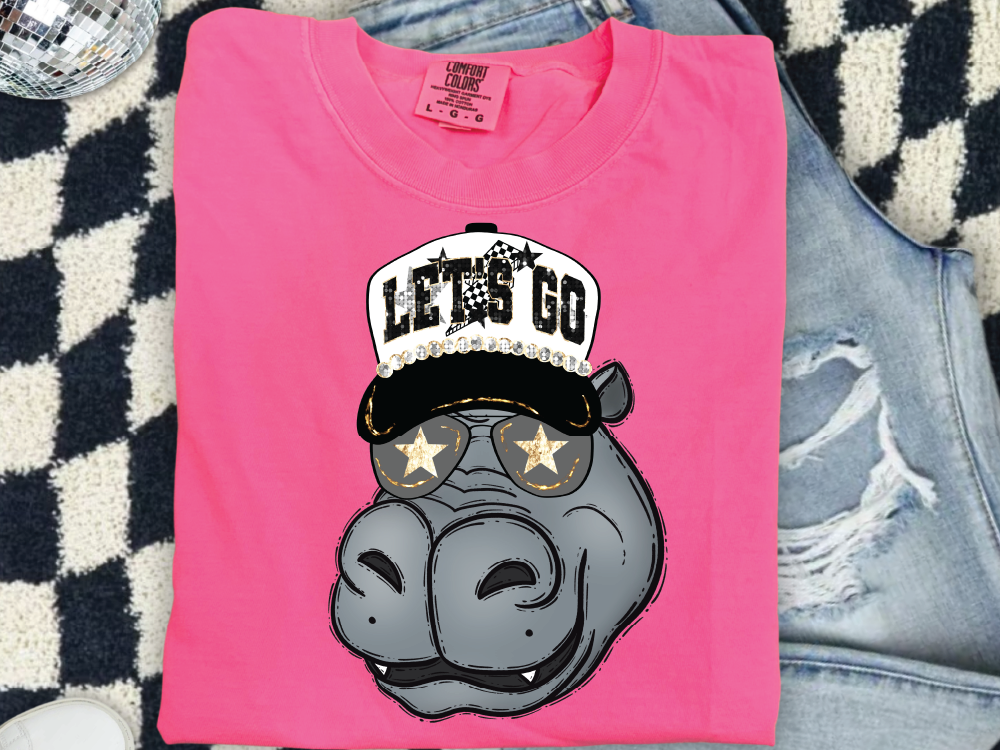 A pink t-shirt with a cartoon image of a hippo wearing sunglasses and a hat with the text "LET'S GO" printed on it.