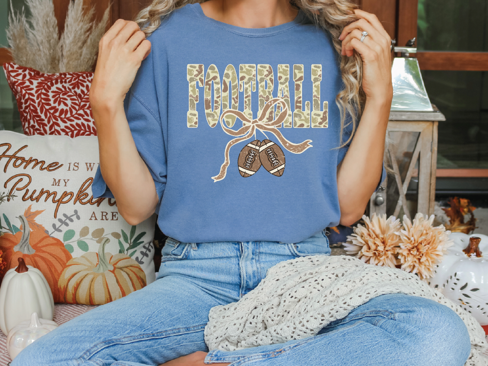 A woman wearing a blue t-shirt with a football graphic and a ribbon, sitting on a couch surrounded by fall decorations.