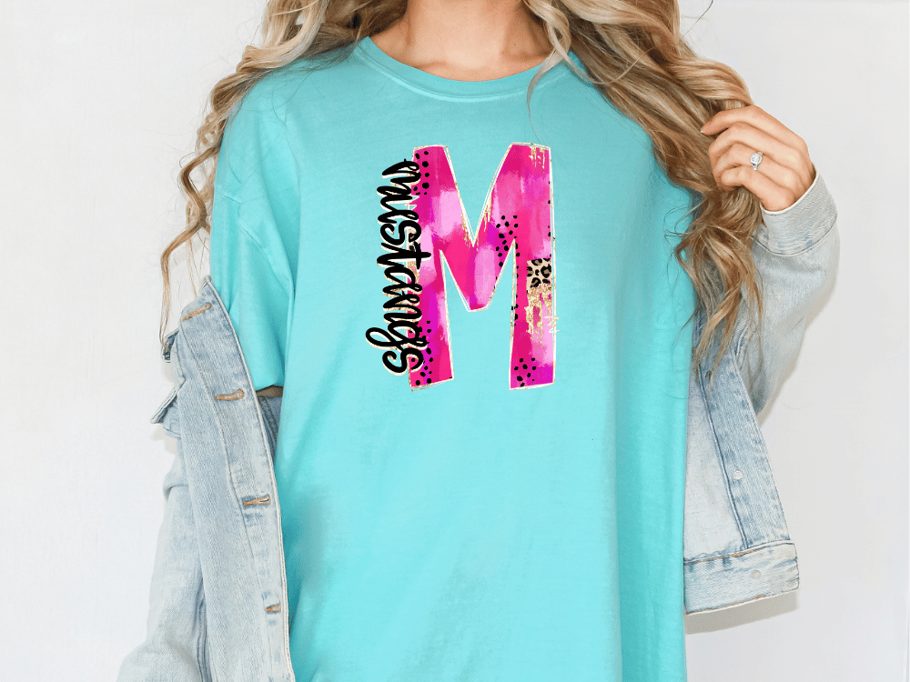 A woman wearing a turquoise t-shirt with a large pink and purple letter "M" printed on it, along with a denim jacket.