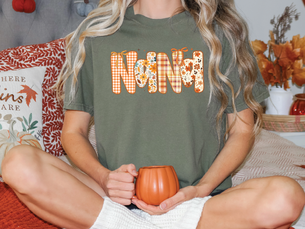 A person sitting on a bed, wearing a green t-shirt with the word "Nana" printed on it, holding a pumpkin-shaped mug.