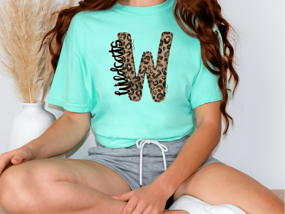 A person wearing a turquoise t-shirt with a leopard print design and gray shorts is sitting on the floor.