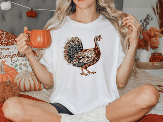 A woman wearing a white t-shirt with a turkey design is sitting on a bed, holding a mug and surrounded by fall decorations.