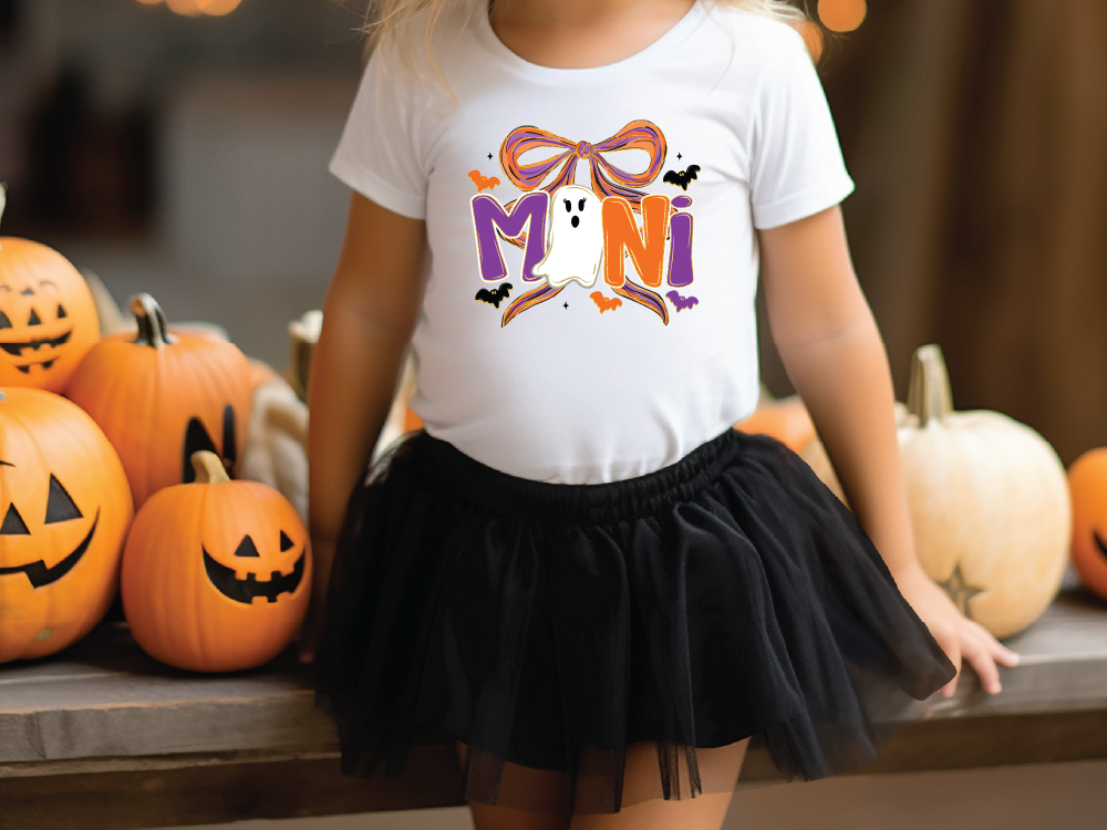 A young girl wearing a white t-shirt with a Halloween-themed design and a black tulle skirt stands in front of a collection of carved pumpkins.