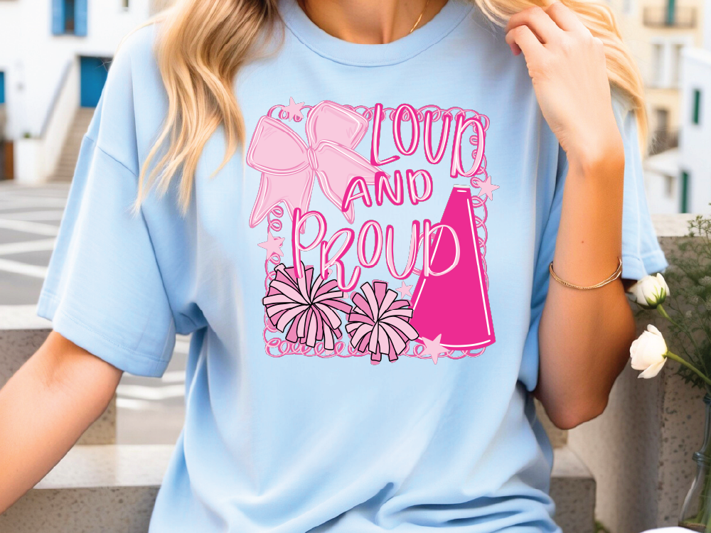 A woman wearing a light blue t-shirt with a graphic design featuring a pink cheerleader, flowers, and the text "LOUD AND PROUD".