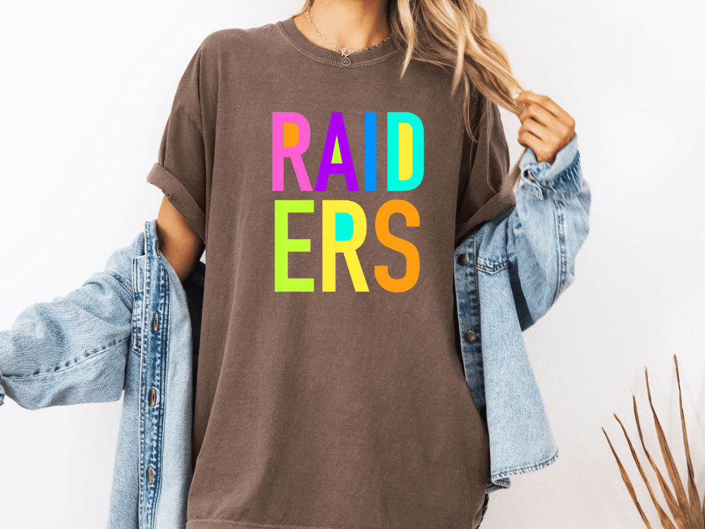 A person wearing a brown t-shirt with the word "RAIDERS" in colorful letters, paired with a denim jacket, stands against a white background.