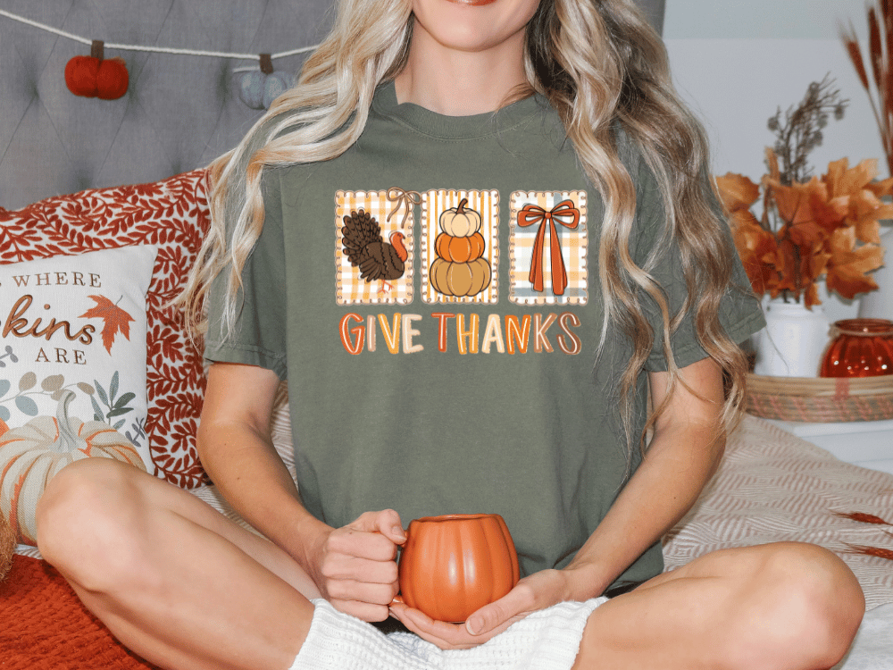 A woman sitting on a bed with a green t-shirt that says "Give Thanks" and holding a pumpkin-shaped mug.