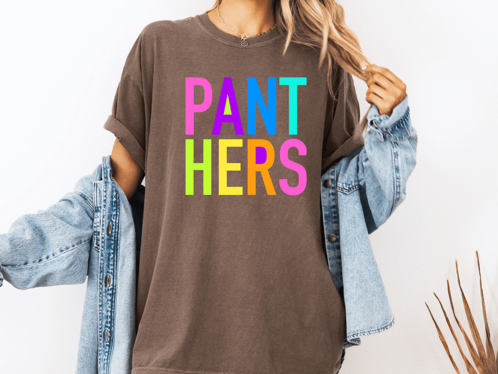 A person wearing a brown t-shirt with the text "PANT HERS" in colorful letters, along with a denim jacket, stands against a white background.