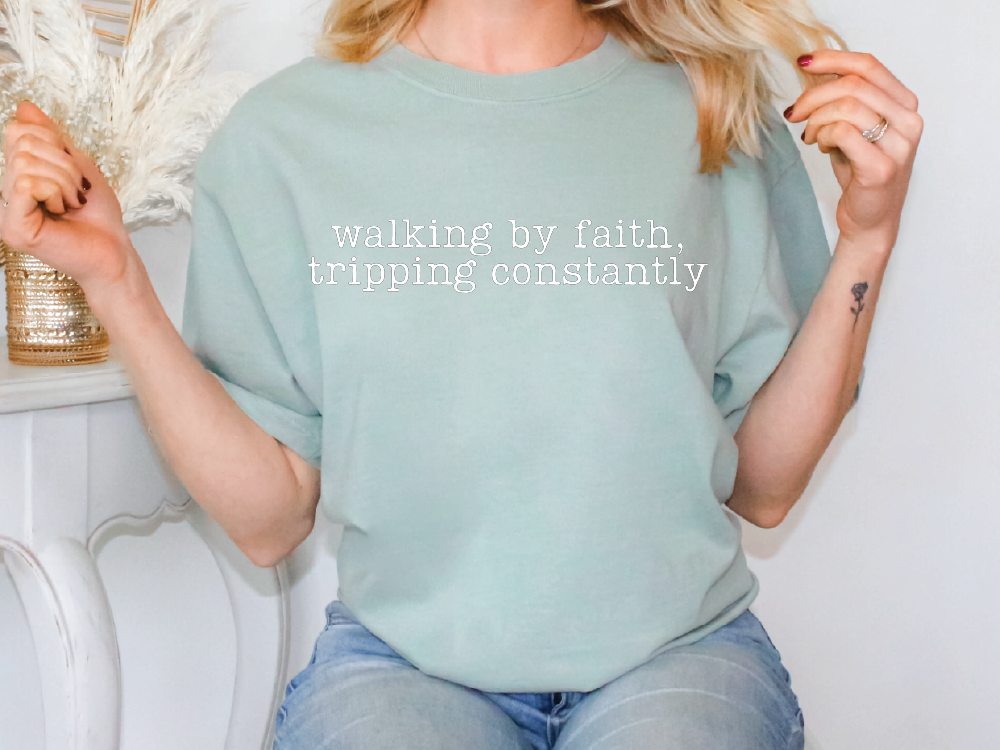 A woman wearing a light green t-shirt with the text "walking by faith, tripping constantly" printed on it.