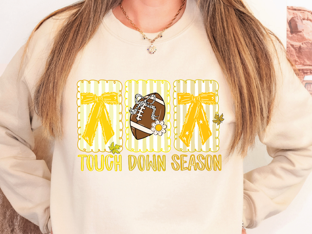 A woman wearing a white sweatshirt with a graphic design featuring a football, yellow bows, and the text "TOUGH DOWN SEASON".