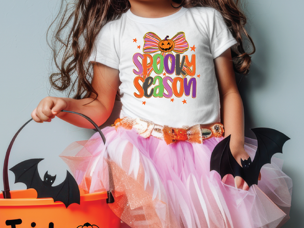 A young girl wearing a white t-shirt with a Halloween-themed design and a pink tulle skirt, holding a black bat and a pumpkin-shaped bucket.