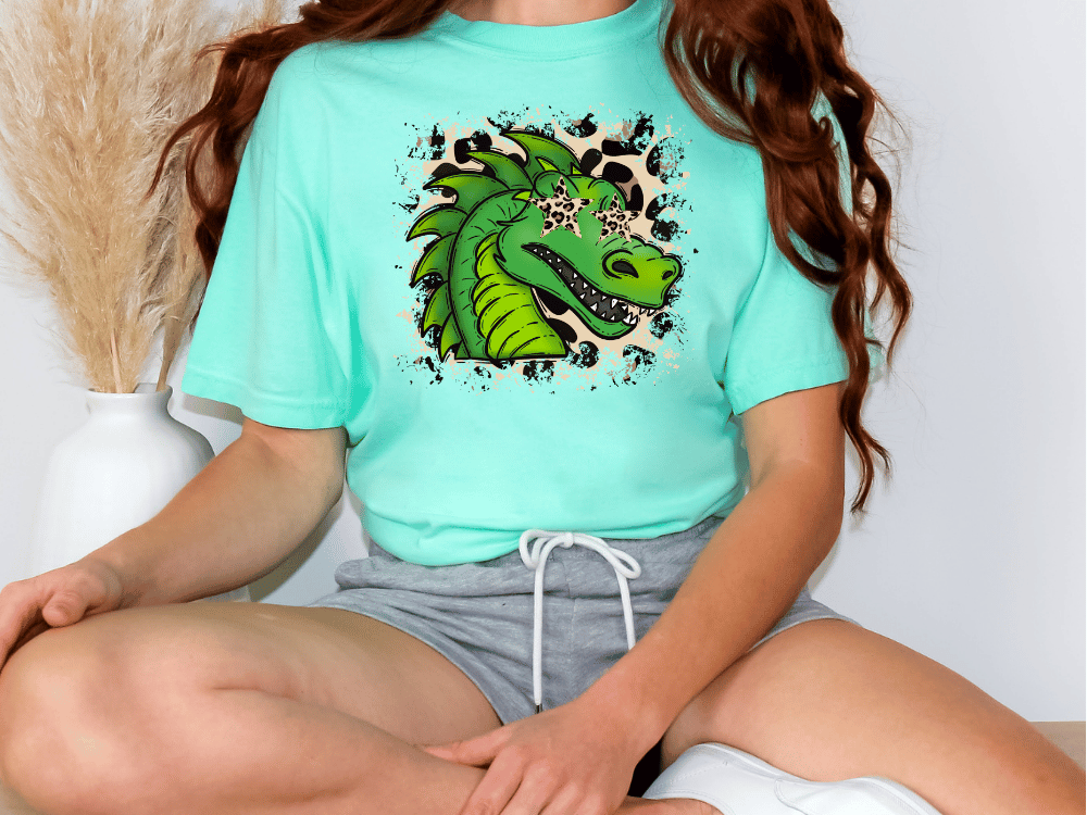 A person wearing a turquoise t-shirt with a graphic of a green dragon and leopard print accents, sitting cross-legged on the floor.