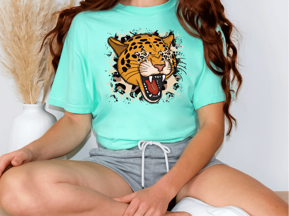 A person wearing a turquoise t-shirt with a graphic of a roaring leopard and gray shorts, sitting on the floor with their legs crossed.